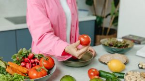 Person in pink overshirt holding a fresh tomato with various organic vegetables on a countertop.